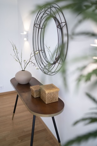 A narrow wooden console table stands against a white wall, adorned with a round decorative wall mirror with a wireframe design. On the table, there is a simple, elegant vase with thin branches and small leaves, alongside two woven baskets. The room has a minimalist, modern aesthetic with a touch of natural elements.