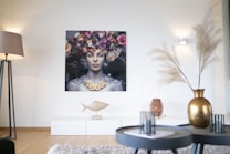 A modern living room features a large artistic portrait of a woman with a floral headdress on the wall. The room is decorated with a minimalist style, showcasing a white sideboard with a sculpture shaped like a fish. Two round coffee tables in the foreground hold candles and a golden vase with dried decorative grass. Soft lighting from a nearby floor lamp and wall sconces create a warm atmosphere.