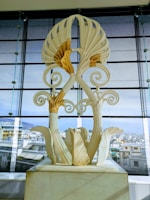 A large, ornate sculpture with intricate floral designs and swirling patterns is displayed indoors with an urban cityscape visible through large glass windows in the background.