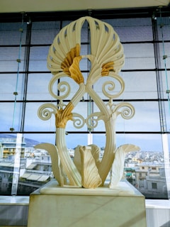 A large, ornate sculpture with intricate floral designs and swirling patterns is displayed indoors with an urban cityscape visible through large glass windows in the background.