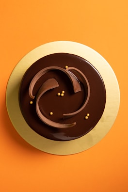 A round chocolate cake with a glossy finish is decorated with elegant swirling chocolate garnishes and small golden spheres. It sits on a golden base, placed against a vibrant orange background.