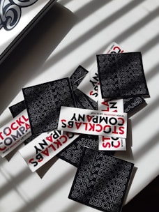 A collection of stickers with bold typography featuring the words 'STOCKLABS' and 'STOCKLABS COMPANY' on a flat surface. The mix includes both black and white stickers, with the black stickers displaying text in a repeating pattern and the white ones featuring red and black text. Sunlight casts shadow patterns across the scene.