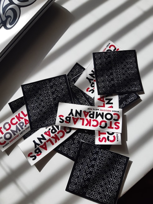 A collection of stickers with bold typography featuring the words 'STOCKLABS' and 'STOCKLABS COMPANY' on a flat surface. The mix includes both black and white stickers, with the black stickers displaying text in a repeating pattern and the white ones featuring red and black text. Sunlight casts shadow patterns across the scene.