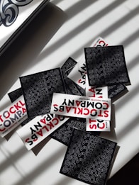 A collection of stickers with bold typography featuring the words 'STOCKLABS' and 'STOCKLABS COMPANY' on a flat surface. The mix includes both black and white stickers, with the black stickers displaying text in a repeating pattern and the white ones featuring red and black text. Sunlight casts shadow patterns across the scene.