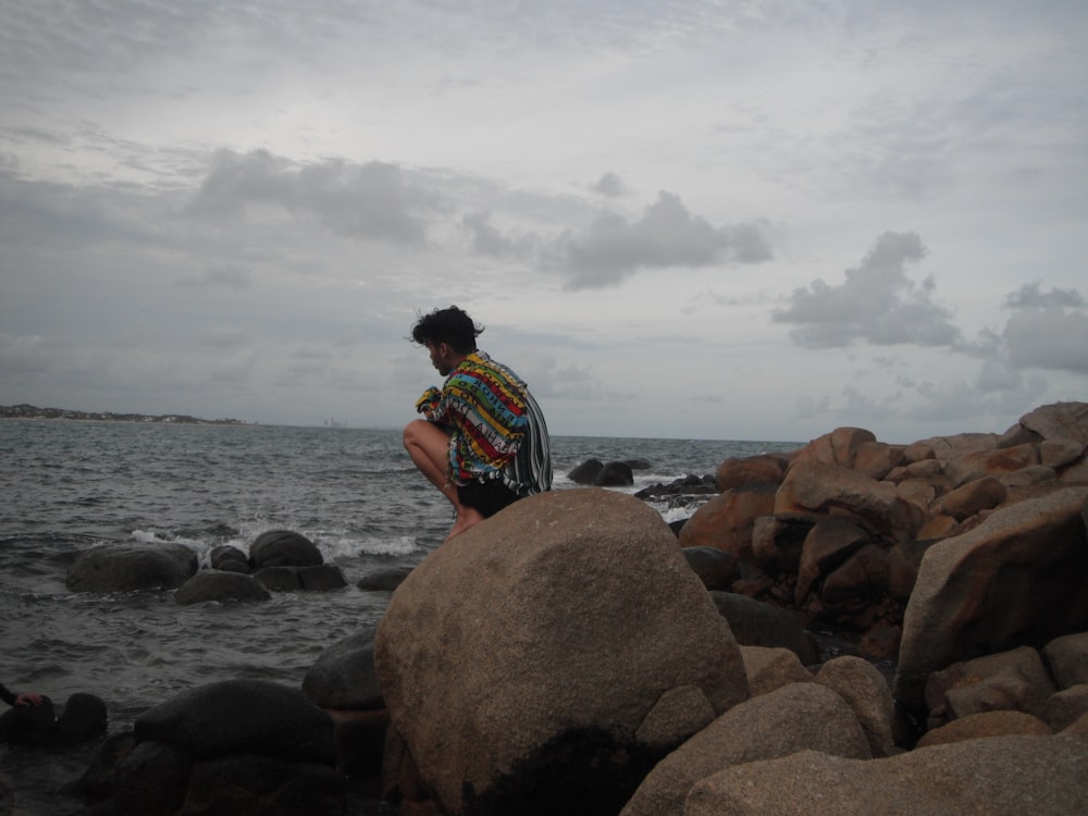 man in blue white and red striped shirt sitting on brown rock near body of water