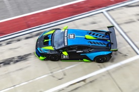 A high-performance sports car in motion on a race track, featuring a sleek design with vibrant blue and green accents. The car has the number 31 on the side and sponsors' names prominently displayed.