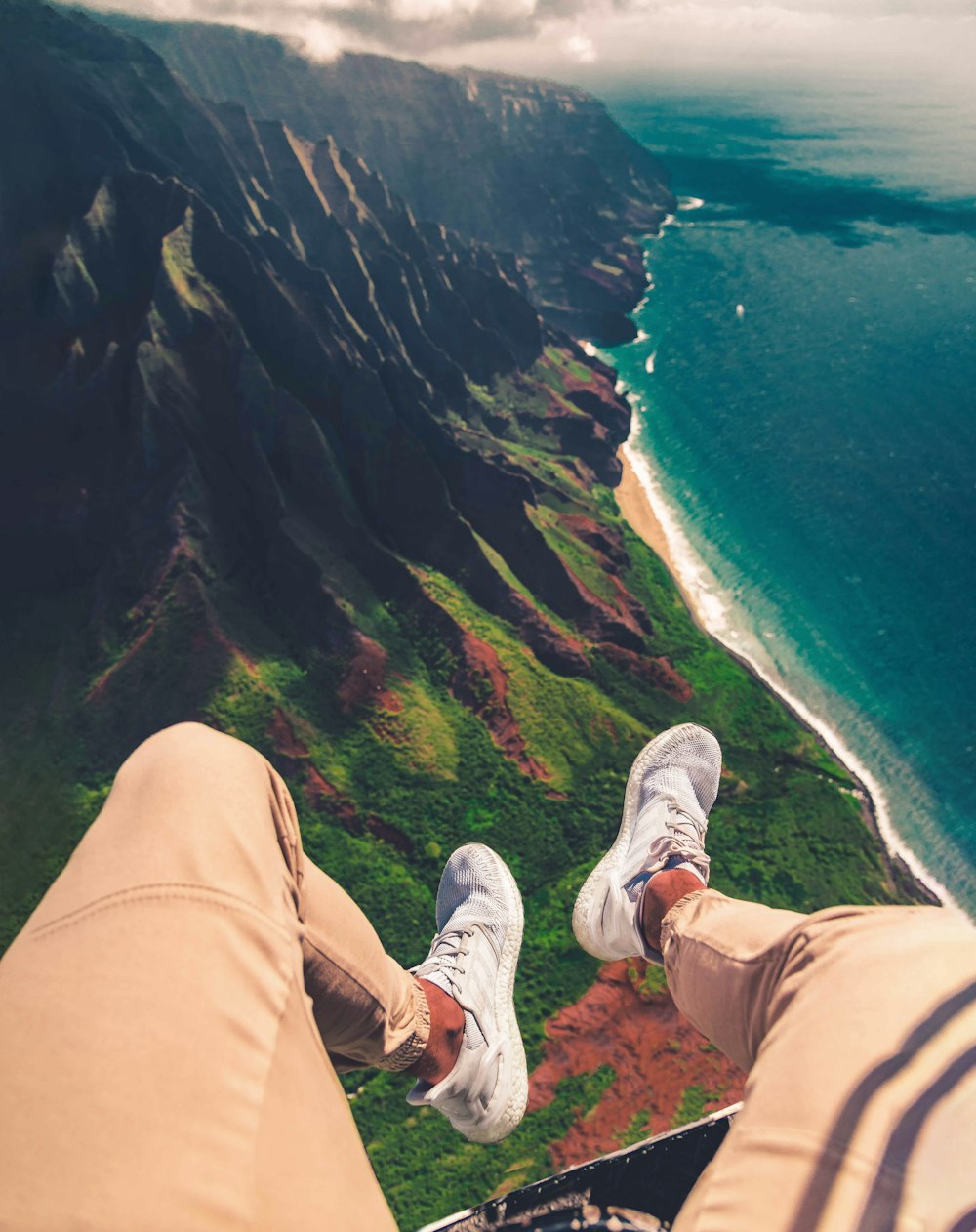 Persona con pantalones marrones y zapatillas blancas sentada en un acantilado sobre el mar