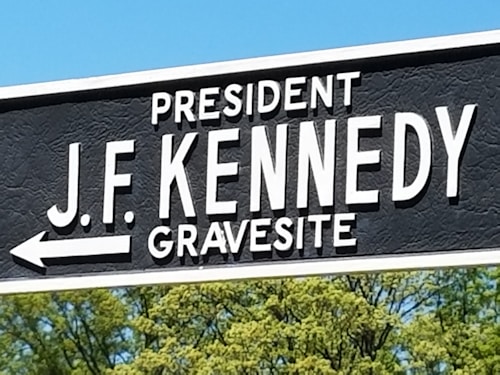 A sign with bold white letters on a black background directs to the gravesite of President J.F. Kennedy. Below the text, a white arrow points to the left. The sign is surrounded by a clear blue sky and framed by the tops of lush green trees.