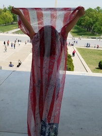 A person stands on a large platform, holding up a translucent fabric with a design resembling the American flag. The background features a busy open area with many people walking around and distant greenery.