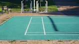 A teal-colored basketball or tennis court with white lines is situated outdoors, surrounded by bricks and some green grass. There are exercise machines in the background, along with a wooden bench to the left.
