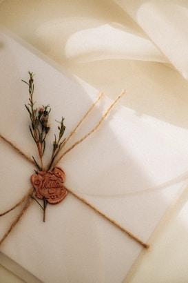 An elegant card is placed on a textured surface, tied with twine forming a cross pattern. A sprig of greenery is tucked under the twine, and a wax seal with intricate details, including the word 'Wedding', is affixed.