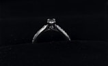 A silver diamond ring is placed on a dark, possibly velvet surface. The ring features a prominent central diamond, with smaller diamonds lining the band on either side.