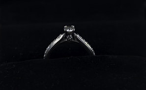 A silver diamond ring is placed on a dark, possibly velvet surface. The ring features a prominent central diamond, with smaller diamonds lining the band on either side.