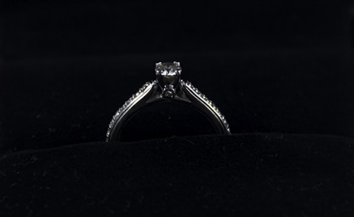 A silver diamond ring is placed on a dark, possibly velvet surface. The ring features a prominent central diamond, with smaller diamonds lining the band on either side.