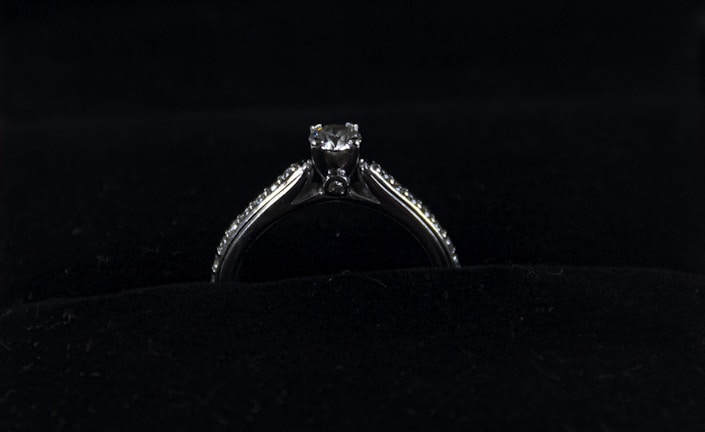 A silver diamond ring is placed on a dark, possibly velvet surface. The ring features a prominent central diamond, with smaller diamonds lining the band on either side.