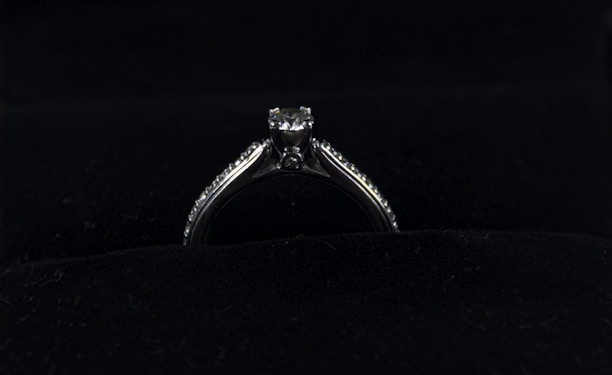 A silver diamond ring is placed on a dark, possibly velvet surface. The ring features a prominent central diamond, with smaller diamonds lining the band on either side.