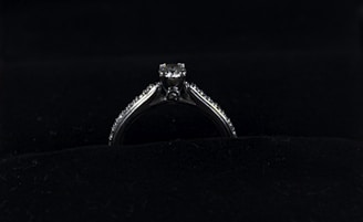 A silver diamond ring is placed on a dark, possibly velvet surface. The ring features a prominent central diamond, with smaller diamonds lining the band on either side.