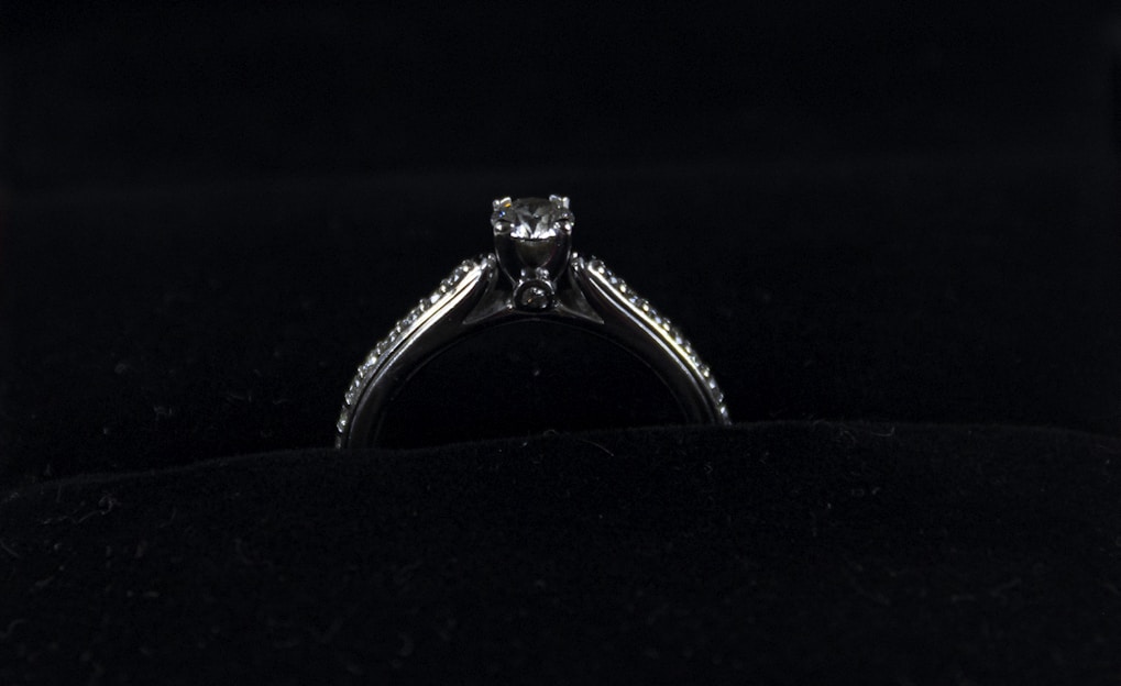 A silver diamond ring is placed on a dark, possibly velvet surface. The ring features a prominent central diamond, with smaller diamonds lining the band on either side.