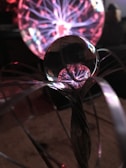 A glass sphere reflects a plasma ball with vivid pink and purple tendrils radiating from its core. The glowing filaments create an electrifying and dynamic pattern of light.