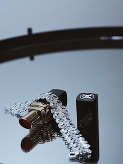 A tube of lipstick with a shiny silver bracelet is elegantly displayed on a reflective surface. The lipstick has a gold casing and a rich red color, while the bracelet features intricate designs and lots of sparkle. A black cap with a logo lies beside the lipstick, adding a touch of luxury.
