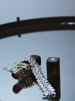 A tube of lipstick with a shiny silver bracelet is elegantly displayed on a reflective surface. The lipstick has a gold casing and a rich red color, while the bracelet features intricate designs and lots of sparkle. A black cap with a logo lies beside the lipstick, adding a touch of luxury.