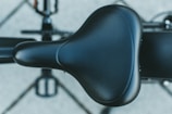 A close-up view of a black bicycle seat with a textured finish, prominently displayed in the center of the image. The bicycle's frame and pedals are visible in the blurred background, suggesting the bicycle is viewed from above.