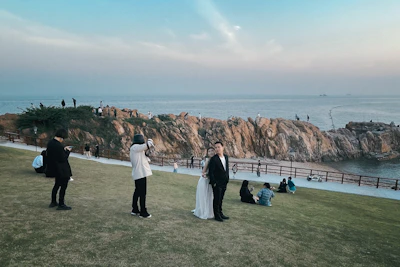 A couple, likely newlyweds, is posing for a photo shoot on a grassy area near rocky cliffs. Several people are seen in the background, some sitting on the grass and others exploring the rocky terrain. The scene overlooks a calm body of water under a mostly clear sky with some light clouds.