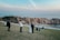 A couple, likely newlyweds, is posing for a photo shoot on a grassy area near rocky cliffs. Several people are seen in the background, some sitting on the grass and others exploring the rocky terrain. The scene overlooks a calm body of water under a mostly clear sky with some light clouds.