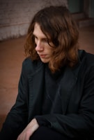 A person with long brown hair is sitting down, looking introspective. They are wearing a black jacket and a dark sweater in an indoor or outdoor environment with a muted, earthy-colored background.