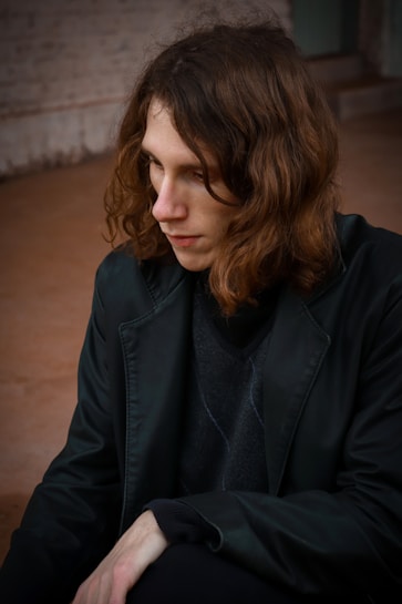 A person with long brown hair is sitting down, looking introspective. They are wearing a black jacket and a dark sweater in an indoor or outdoor environment with a muted, earthy-colored background.