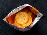 An open bag containing round, crispy snacks with a golden-brown color. The bag is partially made of silver foil on the inside, reflecting the light. The background is a textured dark surface.