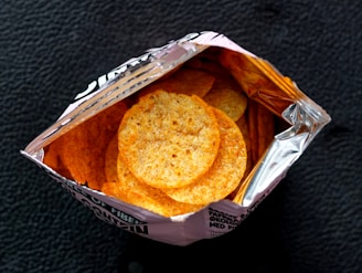 An open bag containing round, crispy snacks with a golden-brown color. The bag is partially made of silver foil on the inside, reflecting the light. The background is a textured dark surface.