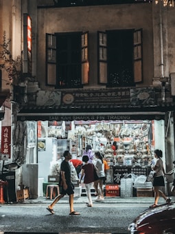 An urban street scene at night with a brightly lit shop that appears to sell various goods, including food items. Several people are walking past or browsing the shop's offerings, which are displayed prominently.
