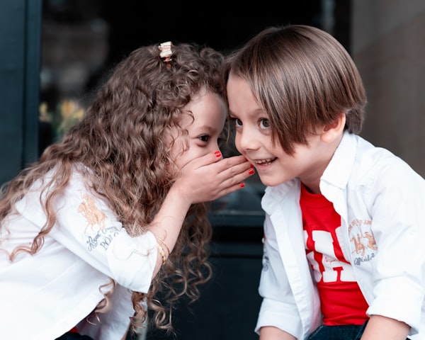 two young kids whispering to each other. Both have a smile on their face.