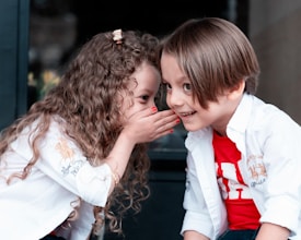 Two young children are sitting closely together, with one whispering into the other’s ear. Both are wearing white shirts, and the child being whispered to has a red shirt underneath. The expressions on their faces suggest sharing secrets or playful communication. The setting appears casual and intimate, possibly indoors.