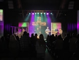 A church service or concert with a band performing on stage. The stage is illuminated with colorful lights and a prominent cross made of lights stands at the center. Musicians are playing instruments, including guitars and drums. The audience is seen sitting and standing, engaged with the performance.