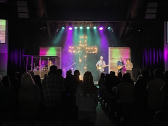 A church service or concert with a band performing on stage. The stage is illuminated with colorful lights and a prominent cross made of lights stands at the center. Musicians are playing instruments, including guitars and drums. The audience is seen sitting and standing, engaged with the performance.