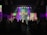 A church service or concert with a band performing on stage. The stage is illuminated with colorful lights and a prominent cross made of lights stands at the center. Musicians are playing instruments, including guitars and drums. The audience is seen sitting and standing, engaged with the performance.