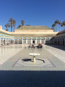 A large, open courtyard with intricate geometric tile patterns on the ground and a central fountain. Surrounding the courtyard is a building with arches and colorful detailing. Palm trees can be seen rising above the building structure. Several people are seated in the distance, adding to the tranquil atmosphere.