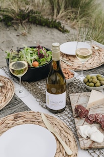 A picnic setup on a beach with a bottle of white wine, two filled wine glasses, a bowl of mixed greens salad with cherry tomatoes, a plate with almonds, a bowl of olives, and a marble board displaying assorted cheeses and slices of salami. The table is decorated with woven placemats and there are plates with metal cutlery. The background features sandy ground with grass tufts.