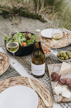 A picnic setup on a beach with a bottle of white wine, two filled wine glasses, a bowl of mixed greens salad with cherry tomatoes, a plate with almonds, a bowl of olives, and a marble board displaying assorted cheeses and slices of salami. The table is decorated with woven placemats and there are plates with metal cutlery. The background features sandy ground with grass tufts.