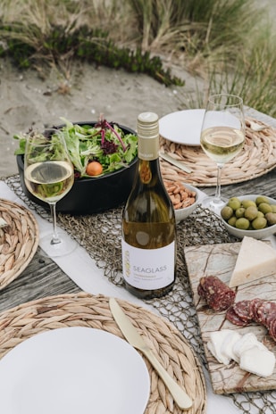 A picnic setup on a beach with a bottle of white wine, two filled wine glasses, a bowl of mixed greens salad with cherry tomatoes, a plate with almonds, a bowl of olives, and a marble board displaying assorted cheeses and slices of salami. The table is decorated with woven placemats and there are plates with metal cutlery. The background features sandy ground with grass tufts.