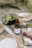 A picnic setup on a beach with a bottle of white wine, two filled wine glasses, a bowl of mixed greens salad with cherry tomatoes, a plate with almonds, a bowl of olives, and a marble board displaying assorted cheeses and slices of salami. The table is decorated with woven placemats and there are plates with metal cutlery. The background features sandy ground with grass tufts.