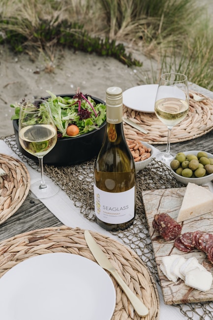 A picnic setup on a beach with a bottle of white wine, two filled wine glasses, a bowl of mixed greens salad with cherry tomatoes, a plate with almonds, a bowl of olives, and a marble board displaying assorted cheeses and slices of salami. The table is decorated with woven placemats and there are plates with metal cutlery. The background features sandy ground with grass tufts.