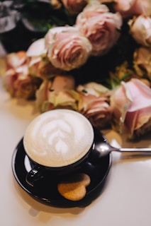 A creamy latte with heart-shaped foam art beside a small vase of fresh flowers.