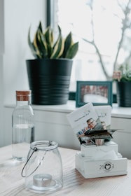 A minimalistic interior surface featuring a wooden tabletop. A glass jar with a tealight candle, a decorative plant in a black pot, and a water bottle with a wooden cap are neatly arranged. Additionally, there is a photo frame in the background and a stand holding printed leaflets.