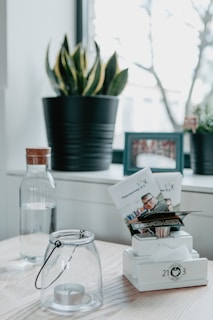 A minimalistic interior surface featuring a wooden tabletop. A glass jar with a tealight candle, a decorative plant in a black pot, and a water bottle with a wooden cap are neatly arranged. Additionally, there is a photo frame in the background and a stand holding printed leaflets.