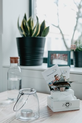 A minimalistic interior surface featuring a wooden tabletop. A glass jar with a tealight candle, a decorative plant in a black pot, and a water bottle with a wooden cap are neatly arranged. Additionally, there is a photo frame in the background and a stand holding printed leaflets.