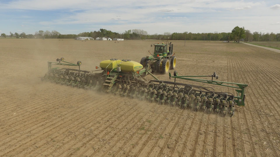 Large tractor pulls yellow-green plow through field with trees in distant background landscape