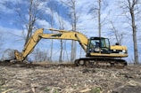 An excavator is actively working on ground excavation in a barren landscape with scattered dry trees and a clear blue sky in the background. The construction equipment is yellow with visible tracks, and a person is operating it from inside the cabin.